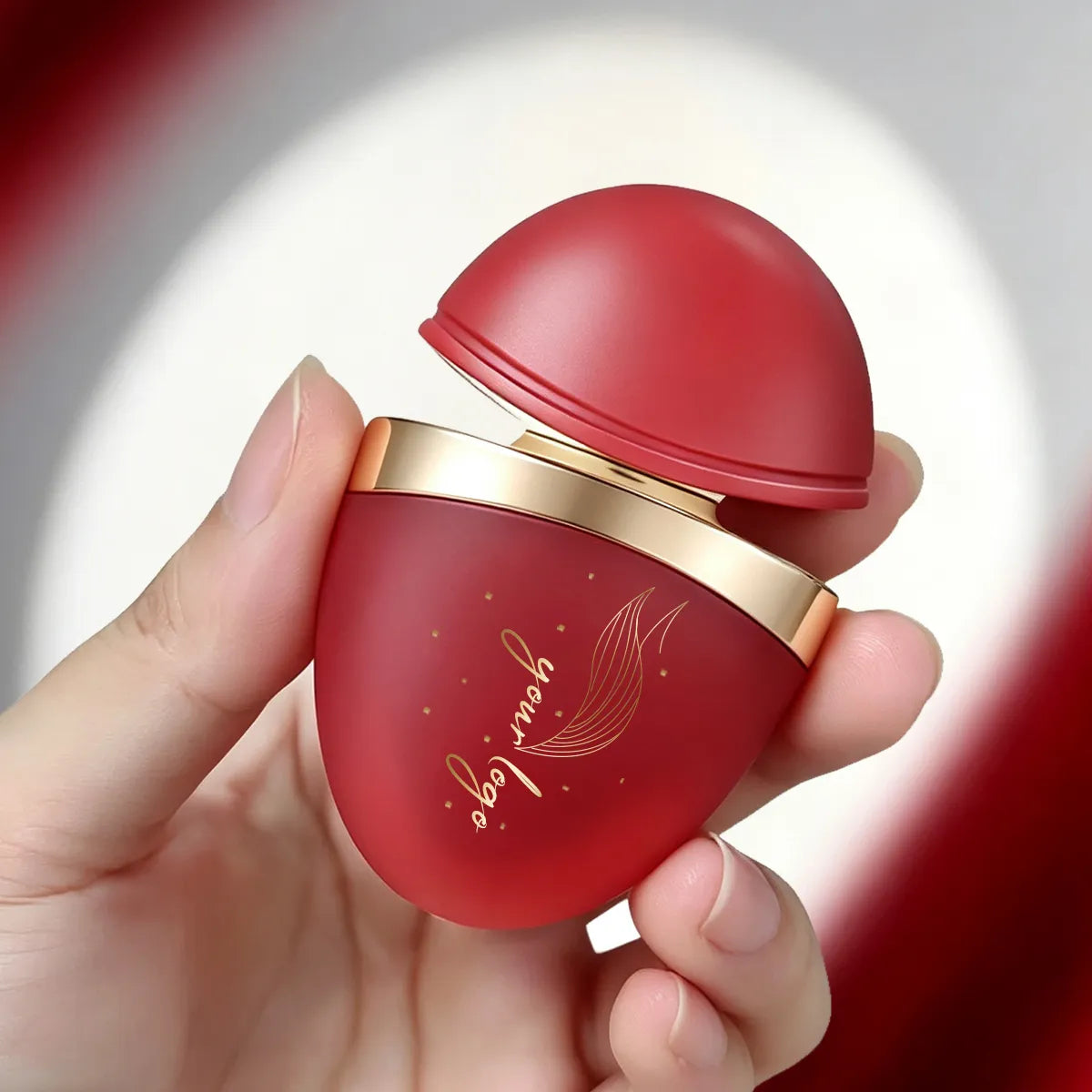 Red fancy lip gloss container held in hand, showing its dome-shaped cap, rounded body and gold-colored neck ring