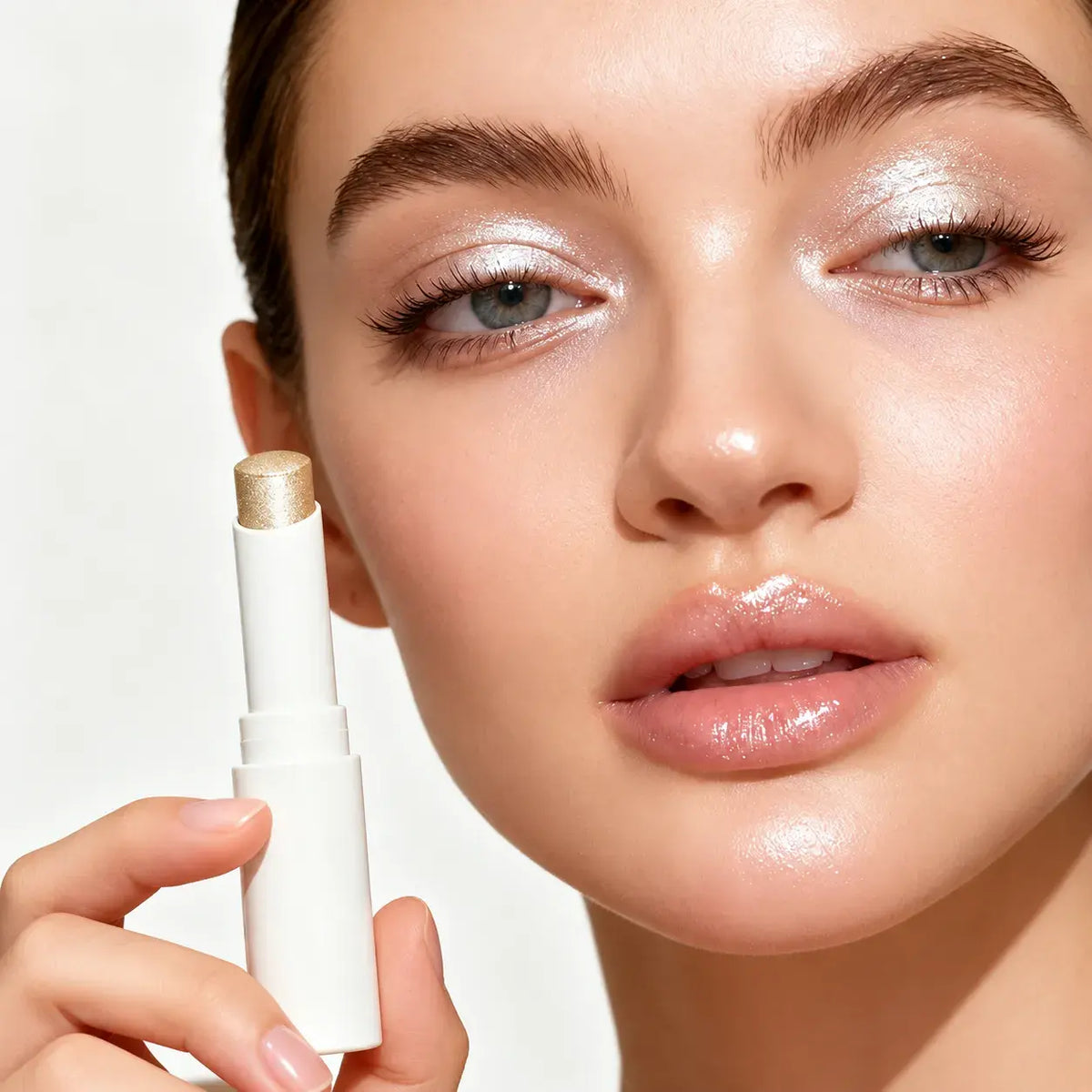 Model holding a white highlighter stick beside her face, with champagne shimmer visible on her cheekbones and eyelids.