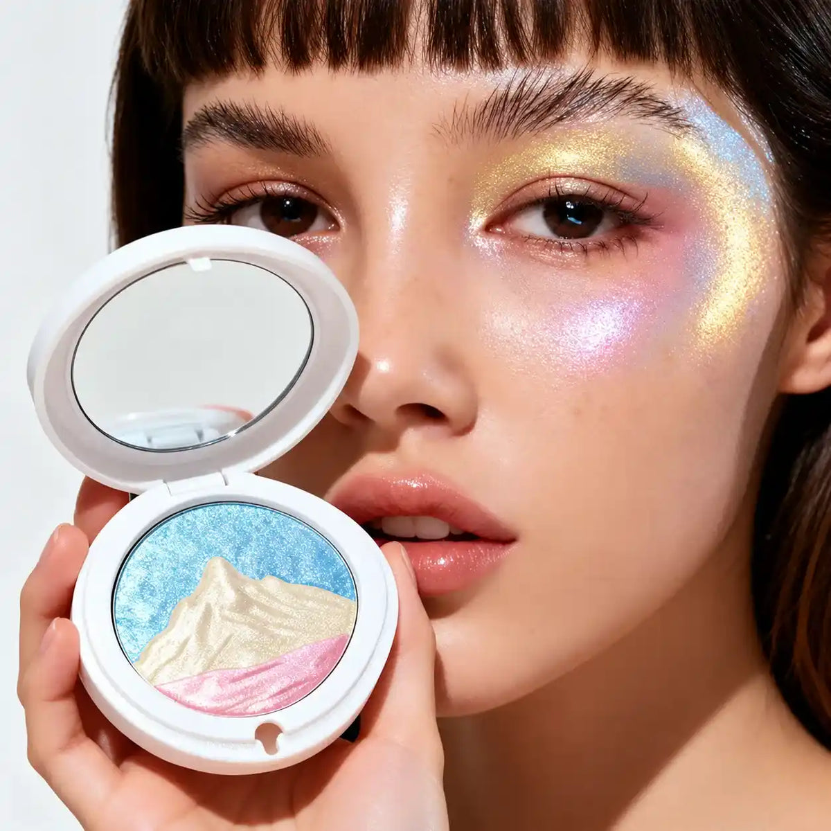 Model holding a three-color putty highlighter with a raised mountain design, showing blue, gold and pink shimmer on her cheek.