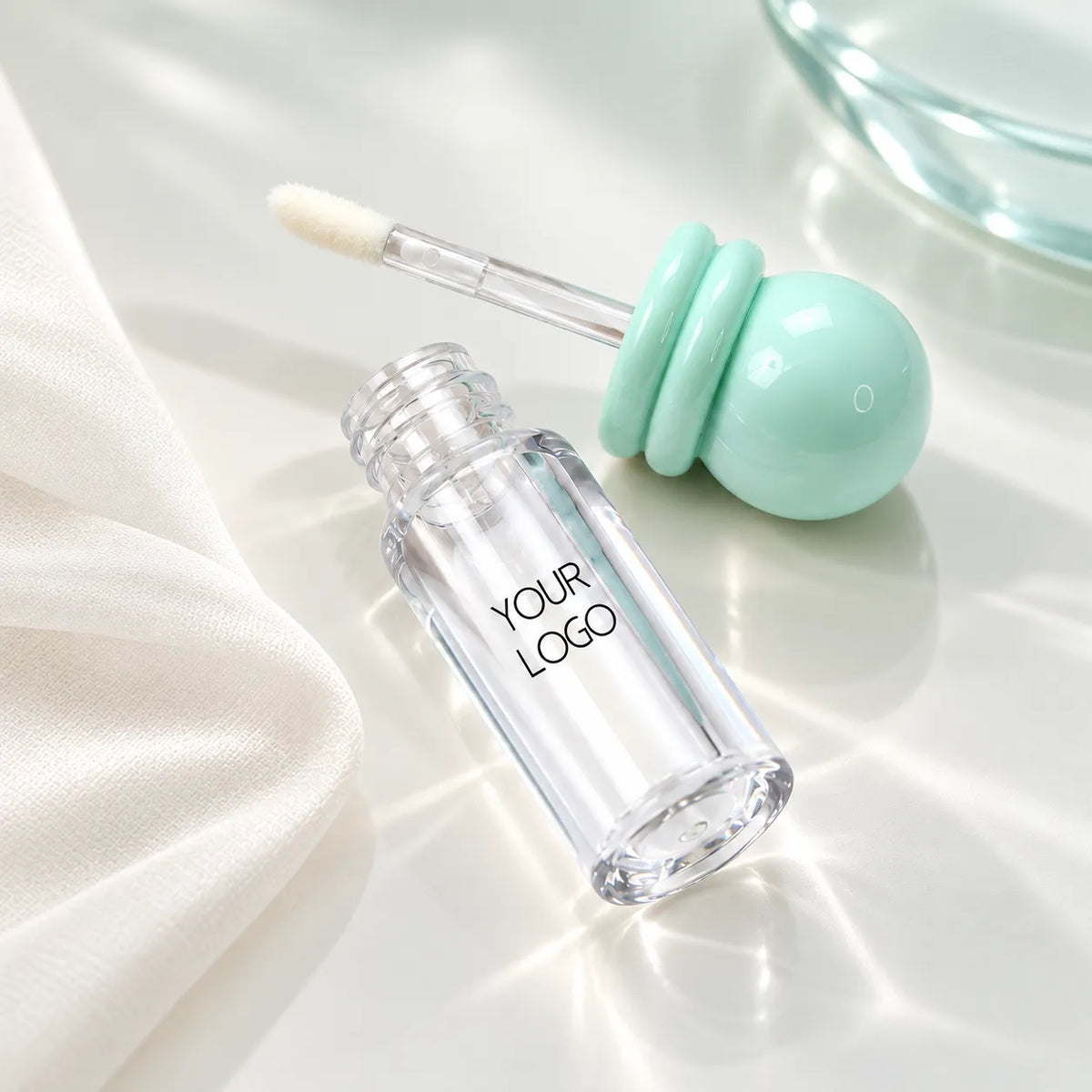 C27100 clear lip gloss bottle beside its mint-green ball-shaped cap with two rounded rings and attached applicator