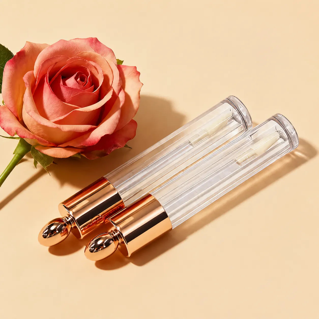 Rose gold empty lip gloss tubes with clear body and rose gold caps displayed beside a rose in an elegant cosmetic packaging scene.